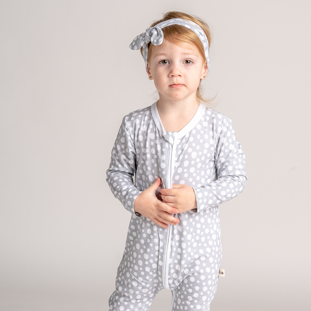 Little girl standing and wearing LiaaBébé sleepsuit in light grey color and top knot headband with dots.