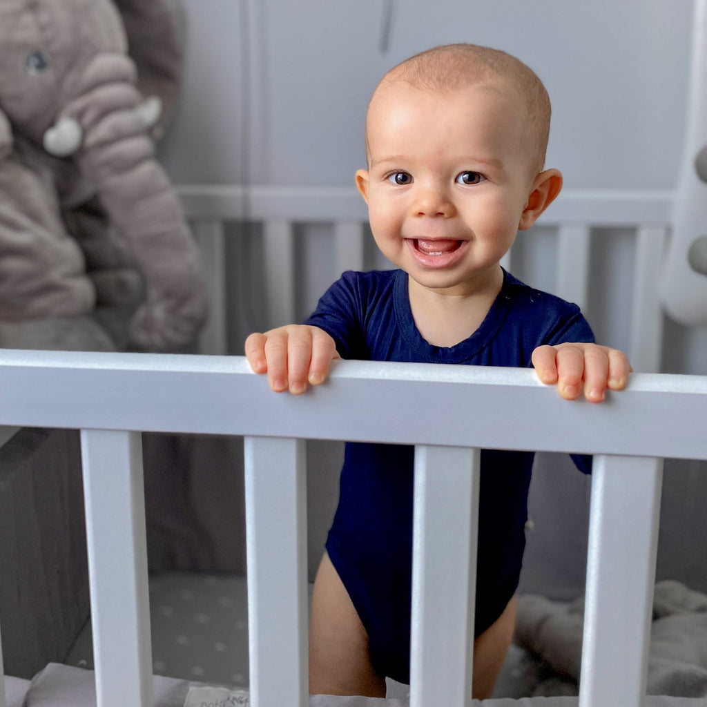 Kisfiú kék bambusz bodyban áll a kiságyban és mosolyog